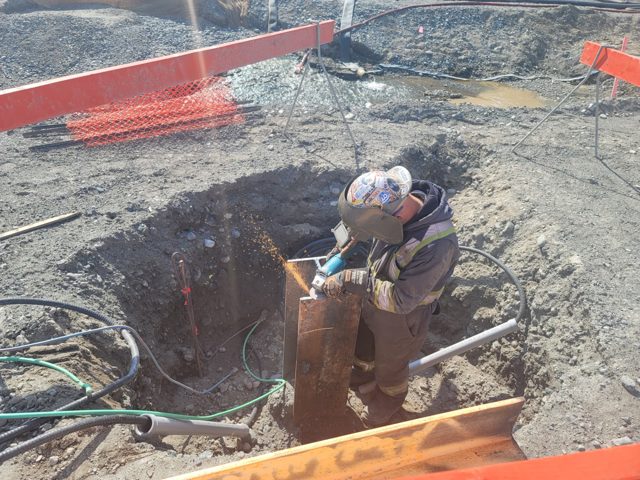 Welder grinding an H-pile cutoff in an open foundation excavation